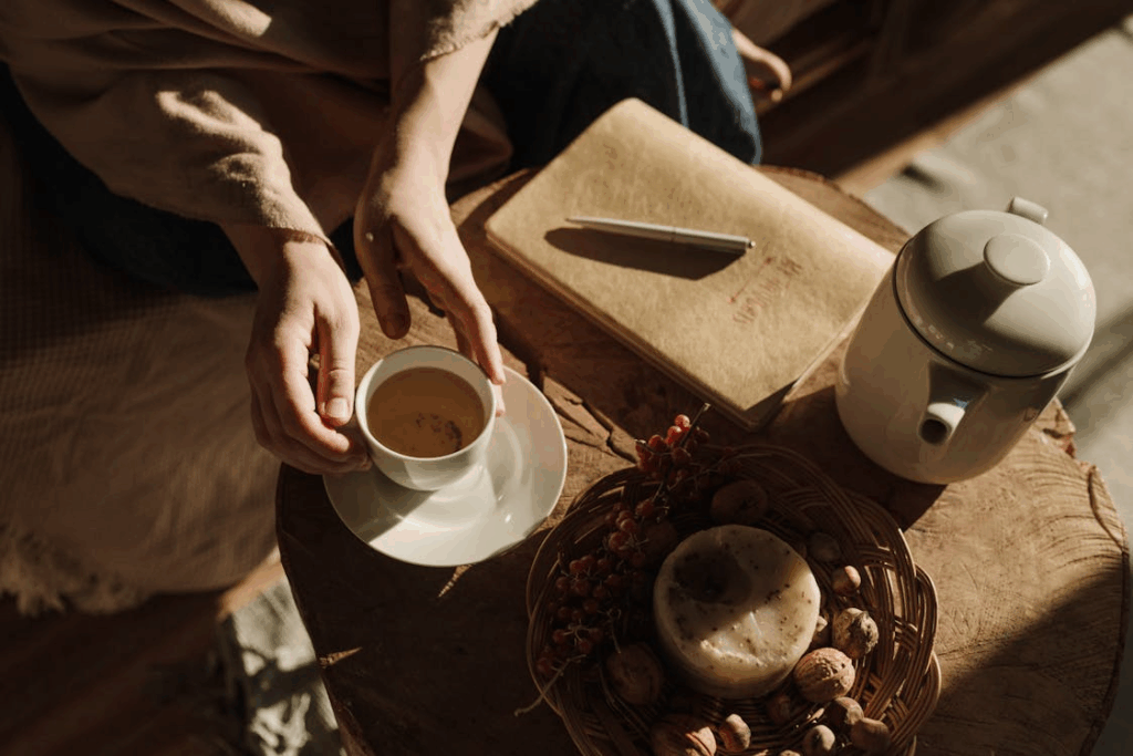 Creator’s hands holding a warm cup of tea beside a journal, capturing a ritual of renewal and reflection.