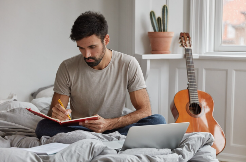 Creative man journaling in a notebook on his bed, capturing ideas for future writing projects.