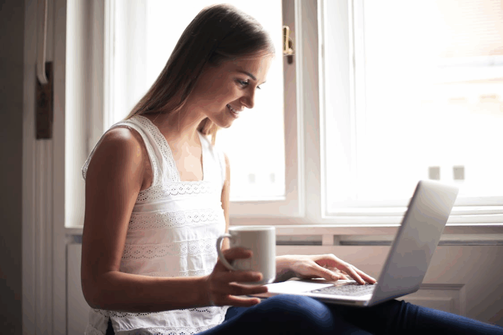 Smiling creator sitting by a window with coffee while sharing her work on a laptop.